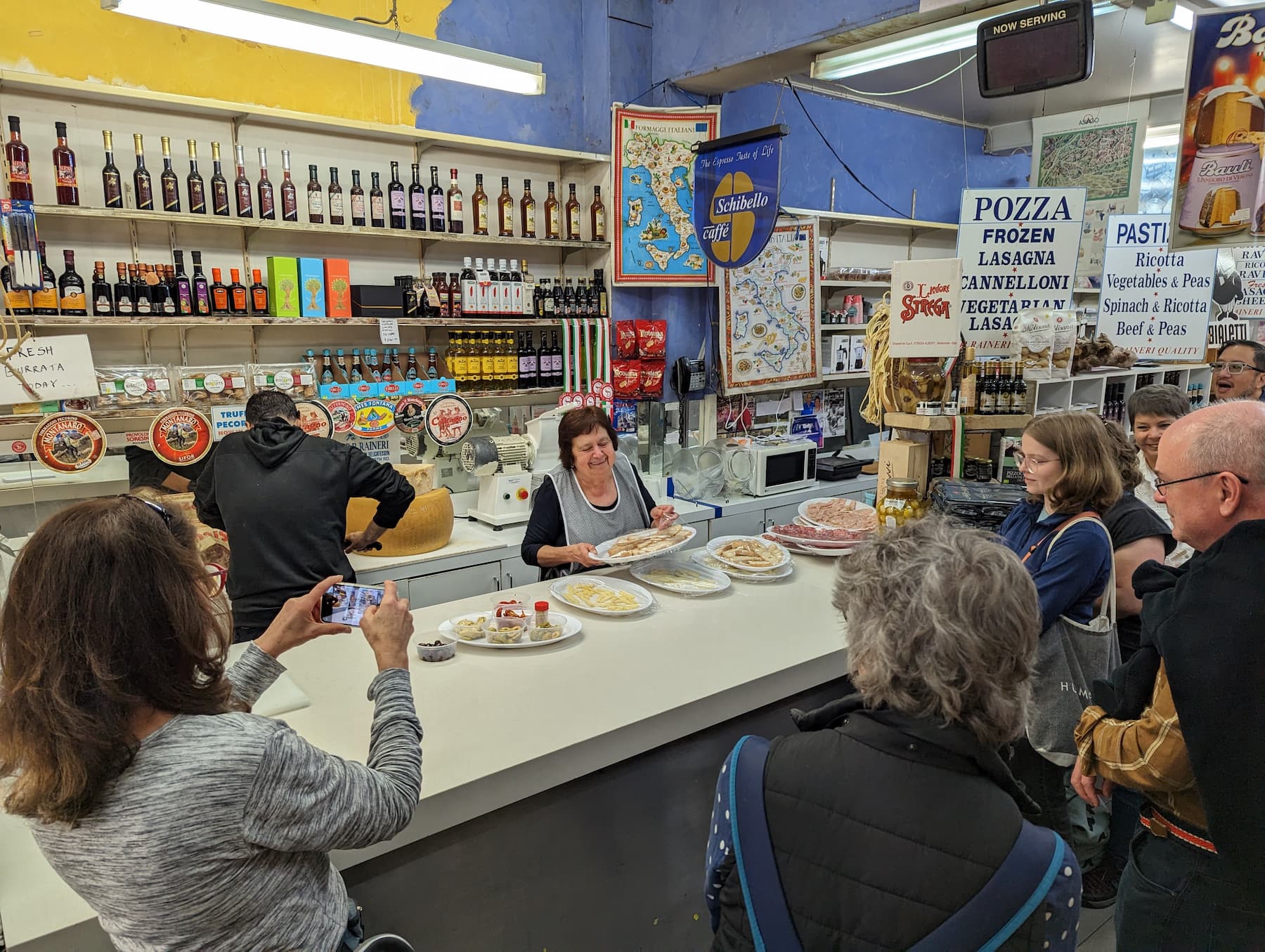 In a small shop in Little Italy, a woman serves various Italian dishes on plates from behind the counter. Several people are gathered around, watching and photographing her. The shelves behind her display numerous bottles and jars, with maps and signs adorning the wall. In a small shop in Little Italy, a woman serves various Italian dishes on plates from behind the counter. Several people are gathered around, watching and photographing her. The shelves behind her display numerous bottles and jars, with maps and signs adorning the wall.